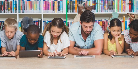 Grundschulklasse mit Tablets auf dem Boden liegend vor Bücherregal